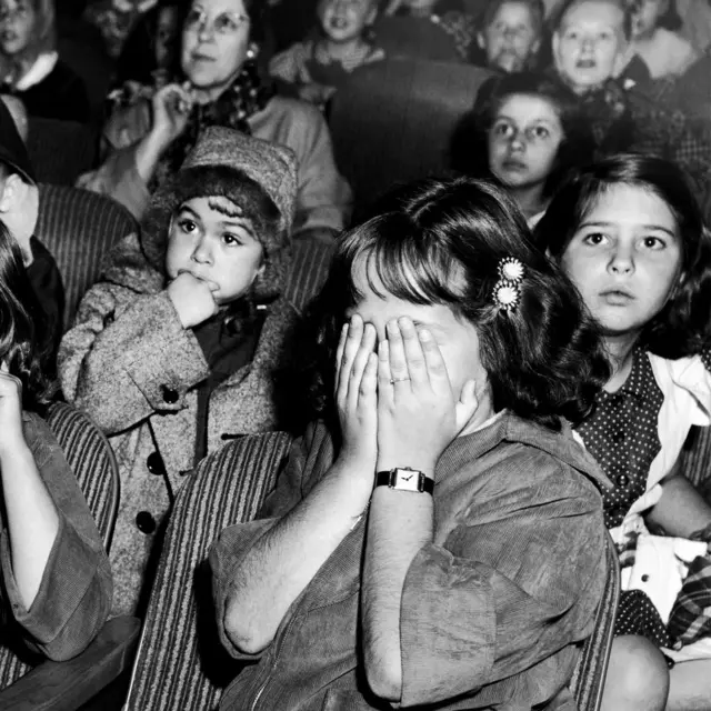 Niñas en una sala de cine viendo una película. Una de las niñas se tapa los ojos, otra tiene la mano sobre la boca y otras más miran con los ojos abiertos de para en par