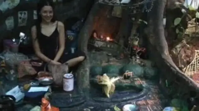 Nina Kutina, en una cueva, junto a las raíces de un árbol y rodeada de objetos decorativos.