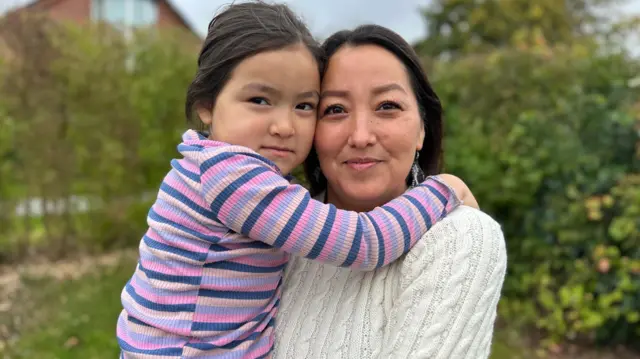 Pilinguaq, vestida con un jersey color crema y pendientes colgantes, sostiene a su hija, que lleva una camiseta de rayas rosas, moradas y azules. La niña abraza a su madre por los hombros, mientras que al fondo se ven árboles y arbustos borrosos frente a una casa.
