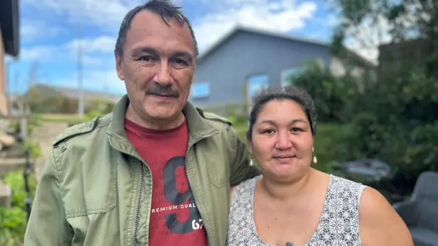 Ulrik y Johanne aparecen en la foto al aire libre. Al fondo se ven un par de edificios, vegetación y un sendero que asciende. Ulrik lleva una chaqueta verde sobre una camiseta roja, mientras que Johanne luce un vestido estampado con escote abierto.