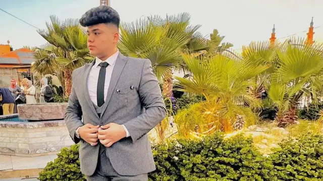 Un joven en un traje gris, camisa blanca y corbata negra, posa frente a unas palmeras en lo que parece ser una celebración solemne