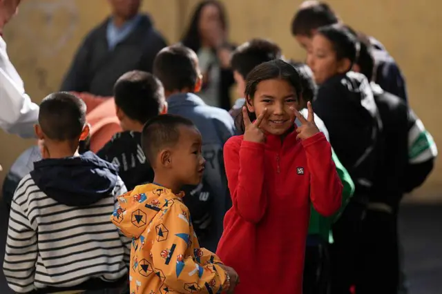 Niños juegan en la calle; en primer plano se ven un niño con una chaqueta naranja estampada y una niña con un suéter rojo que gesticula con ambas manos.
