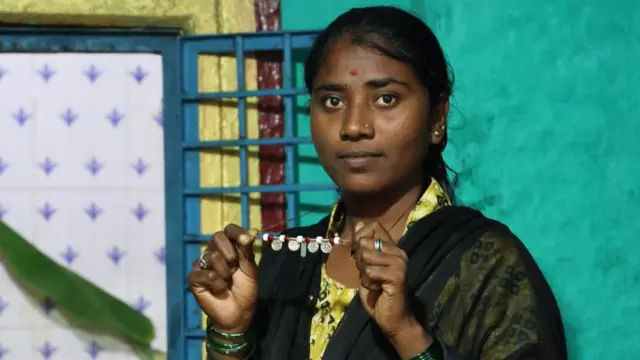 Ankita, una joven devadasi, muestra su collar de perlas. Lleva un vestido amarillo estampado y un chal negro.