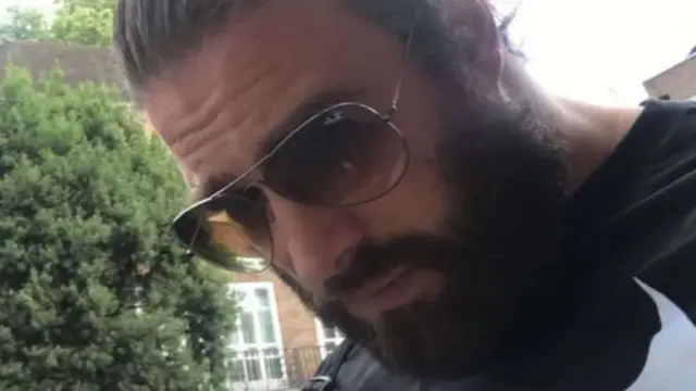 Una foto de Chris Harkins de su perfil de Tinder. Está al aire libre, con árboles y casas al fondo. Lleva el pelo oscuro peinado hacia atrás y barba completa. Usa gafas de sol grandes y tintadas con montura metálica fina. Mira hacia el suelo.