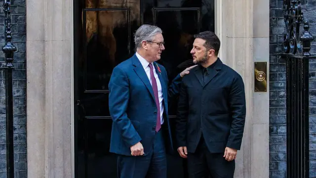 Un hombre con gafas, traje azul, camisa blanca y corbata violeta, le da una palmadita en el hombro a un hombre barbudo con chaqueta y camisa negras en una puerta.