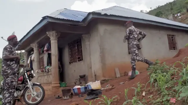Dos policías, uno de ellos en motocicleta, frente a una casa en una colina en Waterloo, Freetown. La casa es de hormigón con techo de chapa ondulada y en el exterior se pueden ver algunas macetas y plantas de maíz.