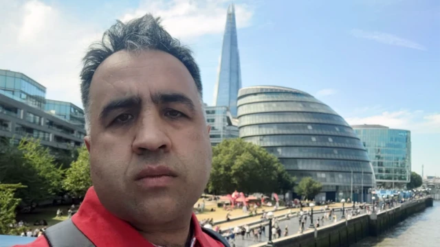 Primer plano de un hombre con cabello corto y oscuro y camisa roja. Está en Londres, con el río Támesis, el antiguo Ayuntamiento y el Shard al fondo.