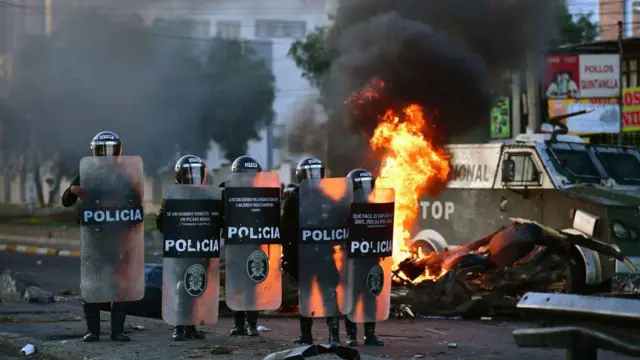 Varios policías bolivianos parados con escudos antimotines frente a un carro en llamas.