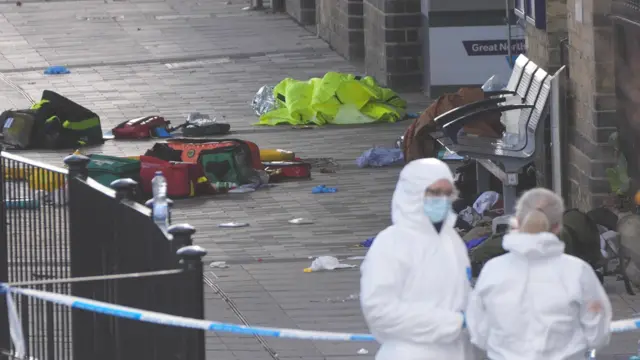 Dos personas, vestidas con trajes de seguridad blancos, se encuentran en un andén. Están detrás de la cinta policial azul y blanca que acordonó una zona llena de basura y equipo de ambulancia.