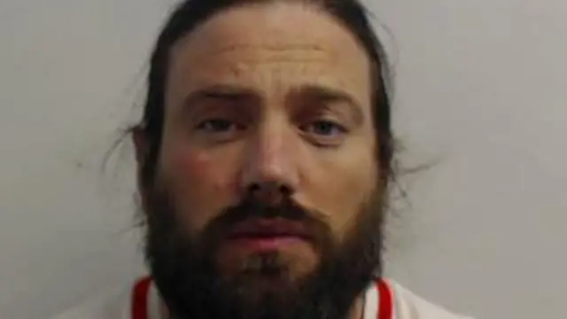 Foto policial de Harkins. Mira fijamente a la cámara. Tiene el pelo largo, recogido, y barba. Tiene aspecto desaliñado.