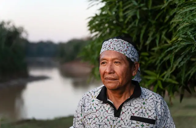 Tomás, con una camisa estampada y un pañuelo a juego alrededor de la cabeza, se encuentra al aire libre, cerca de la orilla de un río. Al fondo se ven aguas tranquilas bordeadas de vegetación,