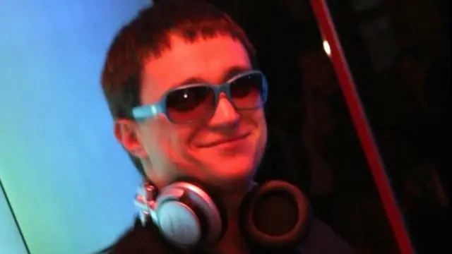 Un hombre con el pelo castaño muy corto lleva gafas de sol y auriculares colgados del cuello. Parece estar dentro de una discoteca.