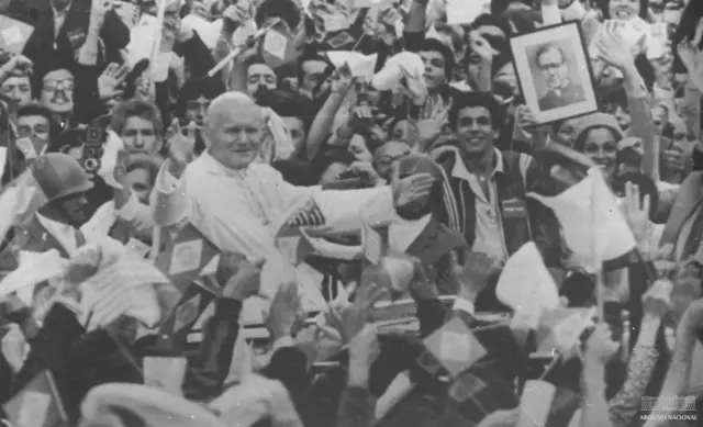 Una fotografía en blanco y negro muestra al Papa en un coche pasando entre una multitud de personas con banderas y carteles brasileños.