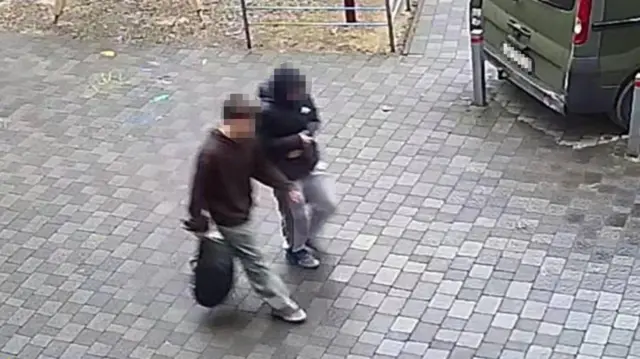 Imagen fija de una cámara de seguridad que muestra desde arriba a dos chicos adolescentes, vestidos de manera informal, caminando por una calle pavimentada. Sus rostros están borrosos. El que está más cerca de la cámara lleva una mochila negra en la mano derecha.