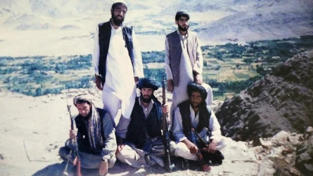Tres hombres están sentados con turbantes y rifles en la mano. Dos hombres están de pie detrás de ellos, vestidos con túnicas, pantalones y sombreros tradicionales de colores claros, y chaquetas oscuras sin mangas. Al fondo se ven campos y montañas.