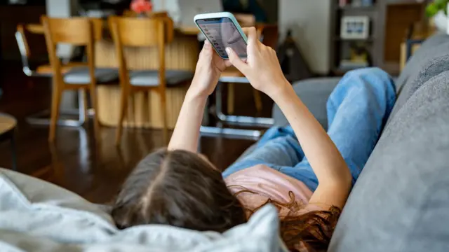 Adolescente en casa mirando las redes sociales en su teléfono móvil mientras está tumbada en el sofá.