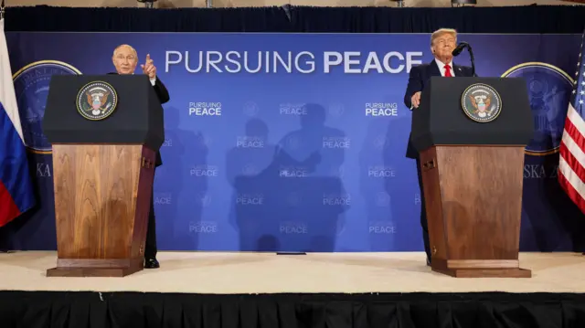Putin habla desde un podio y señala con la mano izquierda mientras Trump está de pie en otro podio a la izquierda de Putin y mira hacia adelante, en Anchorage, Alaska, el 15 de agosto de 2025. Un letrero detrás de ellos dice "En busca de la paz".
