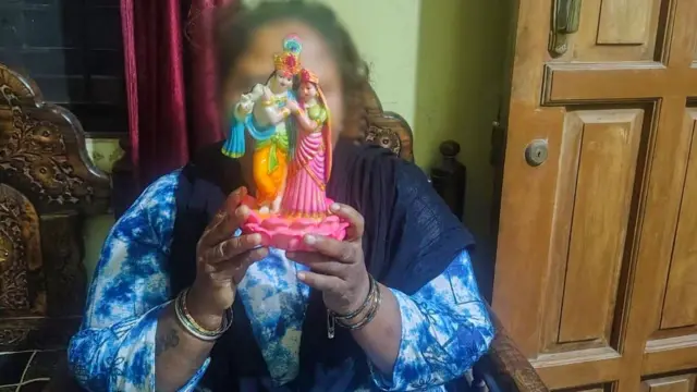Una mujer vestida con ropa azul claro y un chal azul oscuro está sentada en una silla de madera y sostiene la imagen de los ídolos hindúes Radha y Krishna para cubrirse el rostro.
