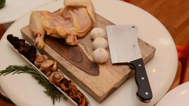 Un cuchillo de carnicero con un mango negro puesto al lado de tres cabezas de ajo y un pollo crudo sobre una tabla 