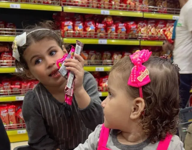 Dos niñas en un carrito de compras, una de ellas sostiene varios paquetes de dulces. Ambas tienen lazos en el pelo. Detrás de ellas están los estantes de un supermercado casi llenos de bolsas con papas fritas y otros productos