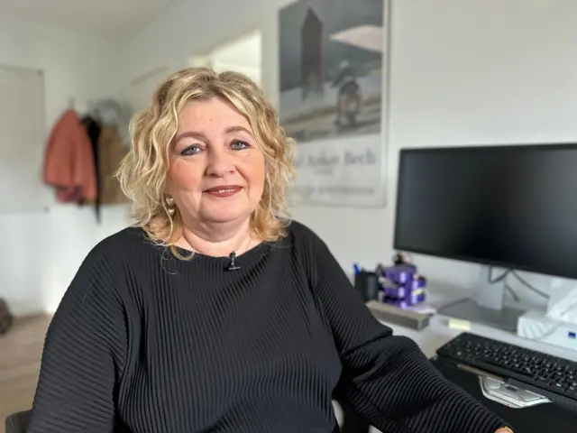 Tordis Jacobsen, una mujer de cabello rubio ondulado hasta los hombros, mira a la cámara. Lleva un suéter negro y está sentada junto a un escritorio con una computadora en una oficina.