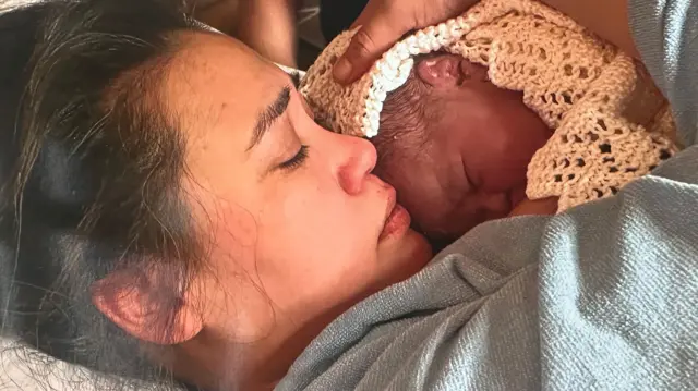 Keira está acostada, acunando contra su pecho a su bebé recién nacida, Zammi, que está cubierta con una manta.