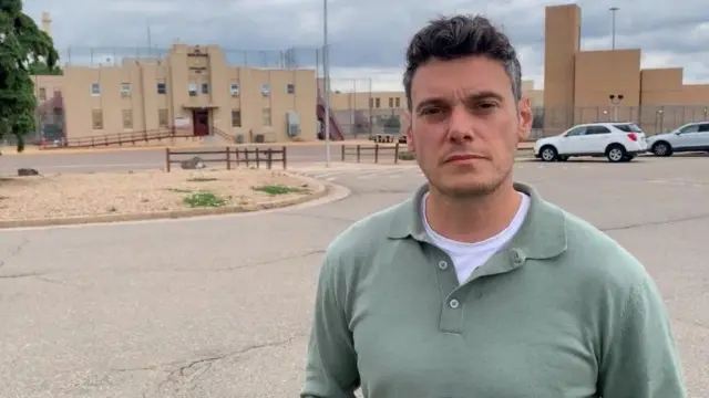 Un hombre con una camisa verde claro está de pie sosteniendo un micrófono frente a un edificio de ladrillo marrón claro rodeado de altas vallas.