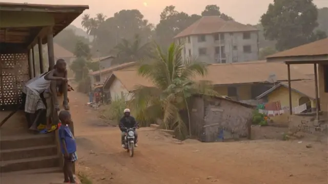 Una motocicleta circula por un camino de tierra en Sierra Leona con niños observando desde un porche.