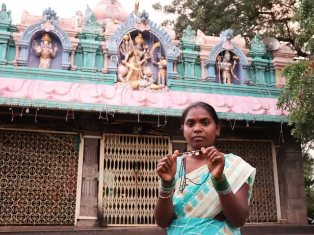 Shilpa está de pie frente a un templo con dioses hindúes decorativos, sosteniendo su collar de cuentas y perlas.