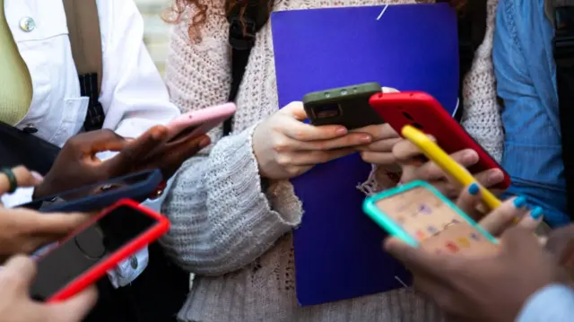 Un círculo de jóvenes desde los hombros hacia abajo sosteniendo teléfonos inteligentes con fundas multicolores.
