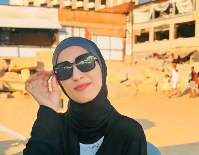 Una mujer con gafas oscuras y un velo negro que le cubre la cabeza está sentada en un asiento en la playa. En el fondo se ven edificios destruidos, uno con las paredes y las ventanas destrozadas