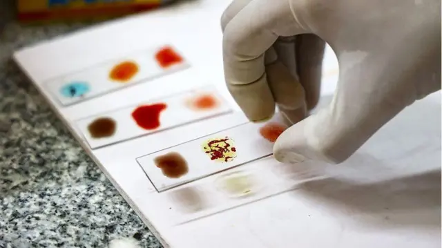 Una mano enguantada sostiene un portaobjetos con gotas de sangre que se están analizando, mostrando muestras con diferentes colores y reacciones.