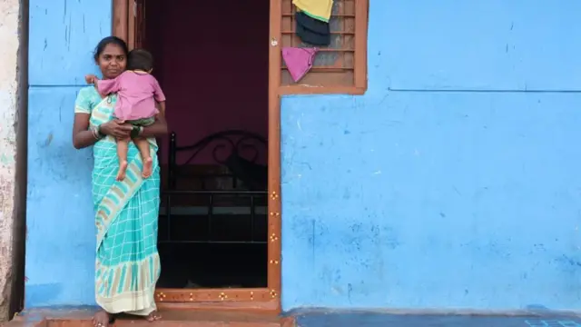 Una mujer india, vestida con un sari verde claro con borde color crema, está de pie frente a la entrada de su casa en el pueblo. Lleva en brazos a su hija de 18 meses.