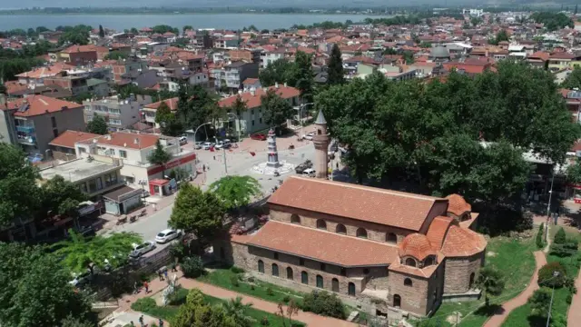 Vista aérea de la Mezquita de Santa Sofía Orhan, un ornamentado edificio de ladrillo con pequeñas cúpulas y un único minarete que da a una pequeña plaza y está rodeado de cuidados jardines. El lago se ve al fondo, al otro lado de la ciudad.