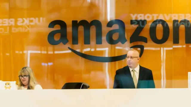 Una mujer sentada frente a lo que parece ser un computador y un hombre en pie, en una oficina de Amazon.