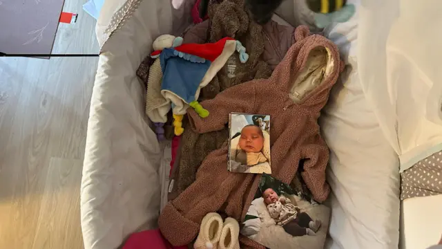 En una habitación del apartamento de Keira se ve una cuna. En el centro de la cuna hay ropa y mantas de bebé, y fotos de Zammi.