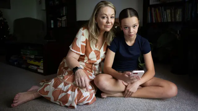 Mel sentada en el suelo con su hija adolescente, Isobel, que sostiene un teléfono rosa.