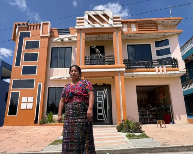 María delante de su casa, un edificio de dos pisos com grandes ventanales, una entrada amplia y de color marrón claro, naranja y rosa pálido