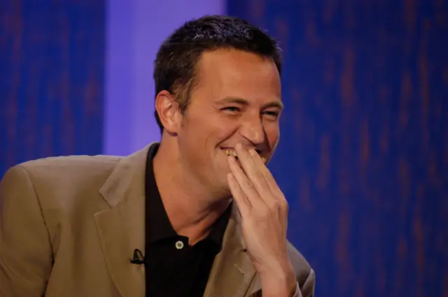 Matthew Perry se cubre la boca mientras ríe. Lleva puesto un blazer color beige y una camiseta tipo polo negra.