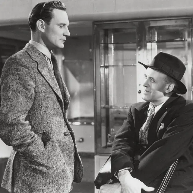 Dos hombres se miran a los ojos. Uno está sentado mientras el otro está de pie frente a él. Ambos llevan traje y corbata. El que está sentado tiene un sombrero. La imagen es en blanco y negro.