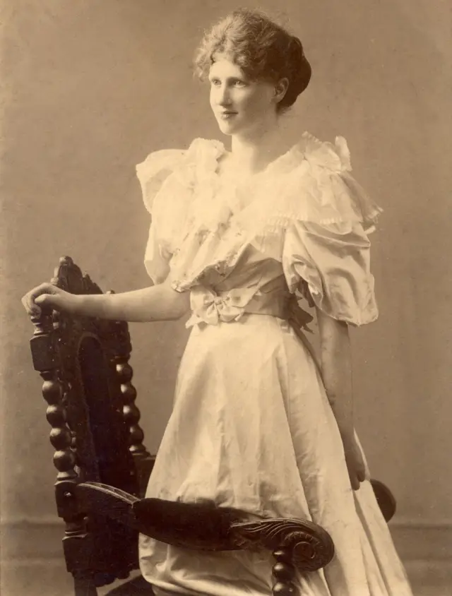 Eglantyne con un vestido largo y claro, apoyada en una silla