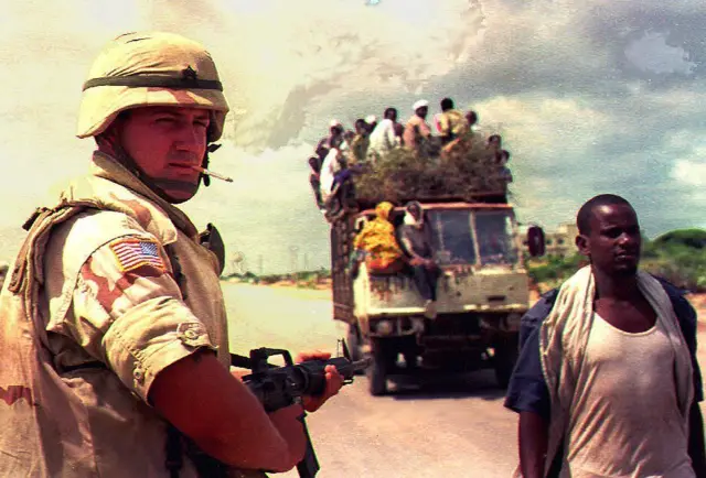 Un soldado estadounidense, con un cigarro entre los labios, vigila una carretera somalí por la que pasan un camión cargado de personas y un peatón.