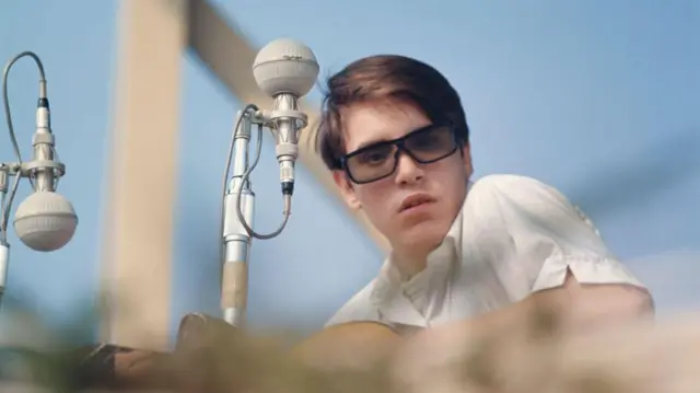 Un joven José Feliciano se presenta en el Newport Folk Festival, en Rhode Island, en 1964. Lleva una camisa de botones blanca, su guitarra en mano y unas gafas oscuras en la cara. Está sentado frente a un micrófono de la época.