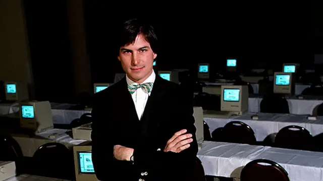 Un hombre con un traje oscuro y una pajarita a rayas verdes y amarillas. Está de pie frente a una fila de computadoras con los brazos cruzados.