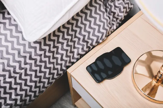 Un teléfono sobre una mesa de noche y al lado la cama.