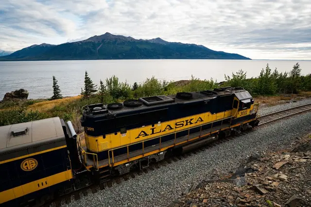 Un tren negro y amarillo con la palabra Alaska escrita en la locomotora, junto a lo que parece ser el mar. Al otro lado se divisa una montaña.