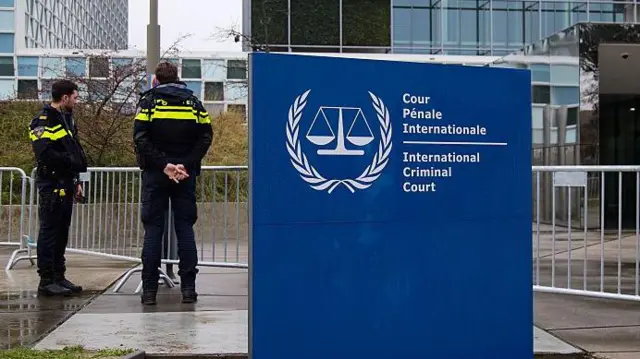 Dos hombres con chaquetas negras y amarillas se encuentran frente a la sede de la Corte Penal Internacional. 