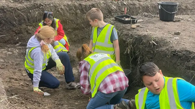 Estudiantes con chalecos reflectantes ayudan en la excavación al oeste de la iglesia de la Abadía de Margam, supervisados por adultos