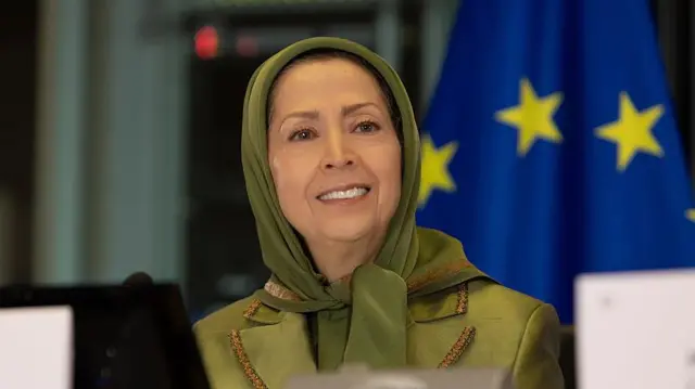 Maryam Rajavi con un pañuelo verde que le cubre la cabeza y una chaqueta del mismo color. Por detrás vemos la bandera de la Unión Europea