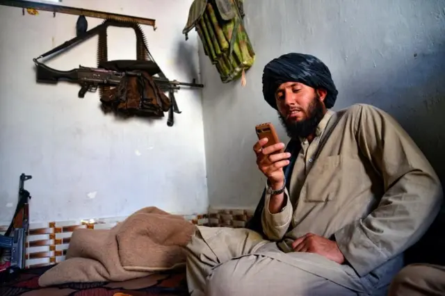Un talibán sentado mirando su celular. Su rifle está colgado en la pared.
